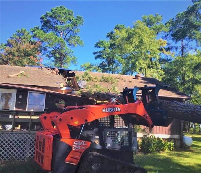 An orange tractor sits in front of a home that had a tree fall on it's roof